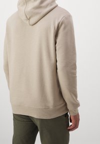 Beige mikina s kapucí vyrobená z texturované bavlněné směsi, která má stahovací kapuci, žebrované manžety a spodní lem, nošená s olivově zelenými kalhotami.