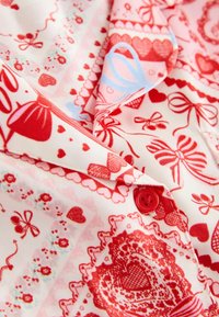 Piros és rózsaszín mintás anyag, melyen szívek, masnik és csipkemotívumok láthatók, piros gombdísszel és lágy, sima tapintású textúrával.