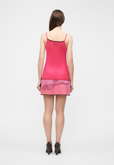 Γυναίκα που φοράει ροζ καμιζόλη και ροζ μίνι φούστα με σχέδια, στέκεται με την πλάτη στην κάμερα σε ένα απλό λευκό φόντο.