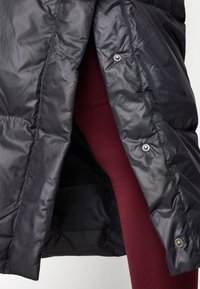 Svart puffjacka med glänsande, kviltad yta delvis öppen, som avslöjar ett par burgundy leggings underneath. Har tryckknappar längs öppningen.