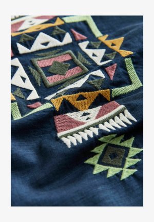 Izšūts tekstils ar tumši zilu fonu un ģeometriskiem rakstiem rozā, zaļā, dzeltenā un krēmkrāsas toņos, izceļot detalizētu šuvumu un tekstūru.