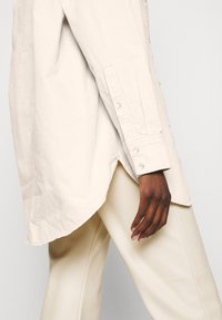 Camicia oversize beige chiaro in morbido tessuto di cotone, con orlo curvo, polsini con bottone e cuciture dettagliate.