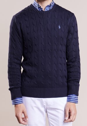 Mann trägt navyblauen pullover mit Zopfstrickmuster über blau-weiß gestreiftem Hemd und weiße Hose, steht vor neutralem Hintergrund.