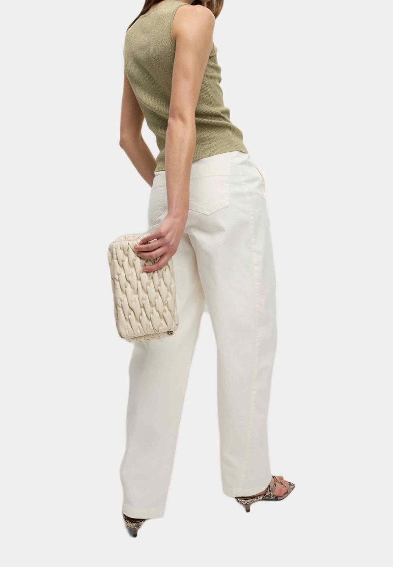 Femme portant un haut vert sans manches et un pantalon blanc, tenant une pochette beige texturée, et portant des talons à motif peau de serpent.