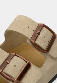 Svetlo bež sandali z dvema širokima trakovoma iz teksturiranega materiala, ki imajo zaponke v bakreni barvi, ter mehkim rjavim notranjim oblogo.