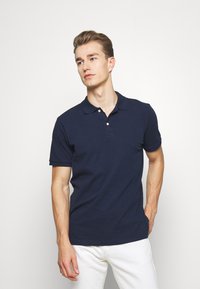 Navy polo shirt made of cotton with a classic collar, two-button placket, and short sleeves. Worn with white trousers.