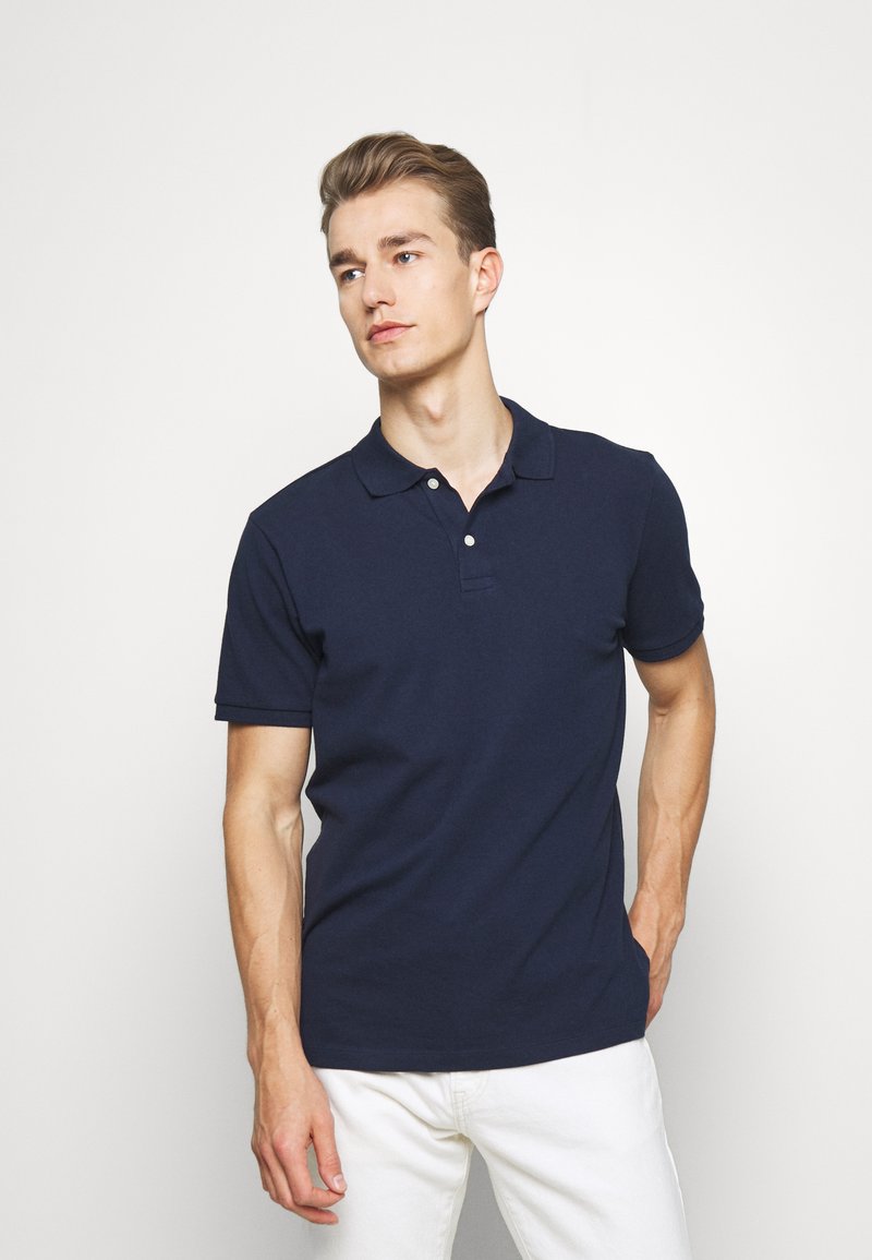 Navy polo shirt made of cotton with a classic collar, two-button placket, and short sleeves. Worn with white trousers.