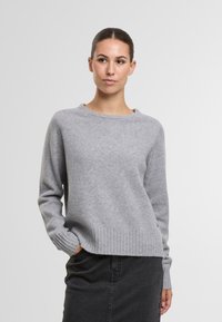 Hellgrauer Strickpullover mit rundem Ausschnitt, gerippten Bündchen und Saum. Weiche Textur; figurbetonter Schnitt, kombiniert mit einem dunklen Rock.