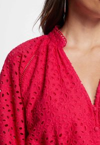 Blusa roja bordada con cuello alto y patrones de ojales intrincados, con un escote en V profundo y una tela suave y texturizada.
