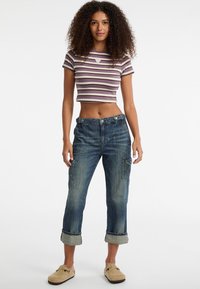 Młoda kobieta z kręconymi włosami, ubrana w fioletową, prążkowaną bluzkę na krótki rękaw, zwinięte dżinsowe spodnie cargo i beżowe buty sportowe, stojąca.