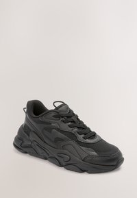 Baskets de sport noires avec semelle épaisse, dessus en mesh et cuir, et fermeture à lacets, montrées sur un fond neutre.