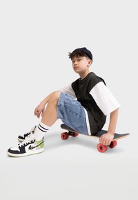 Skateboard s červenými koly, na kterém je osoba v černo-bílém oversized tričku, džínových šortkách a botách s zelenými akcenty.