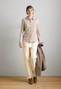 Bež košulja dugih rukava sa dugmadima, krem pantalone širokih nogavica, smeđi gležnjači i smeđi kaput u ruci, postavljeni na neutralnoj pozadini.