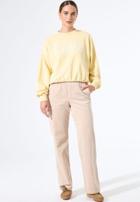 Mujer vistiendo un sudadera amarilla con el texto "El futuro es amor", pantalones de pana beige y zapatos mocasín marrones, de pie con una mano en el bolsillo.