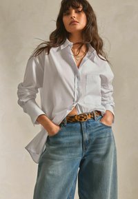 Chemise blanche à boutons avec manches longues, associée à un jean large bleu clair et une ceinture à motif léopard. Un collier en chaîne dorée est visible.
