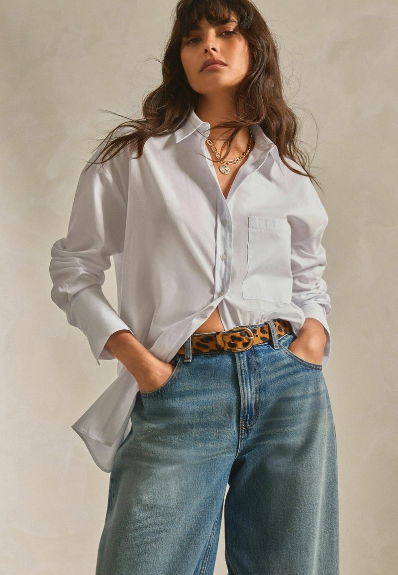Chemise blanche à boutons avec manches longues, associée à un jean large bleu clair et une ceinture à motif léopard. Un collier en chaîne dorée est visible.