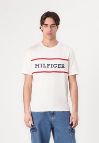 White cotton t-shirt with round neck, featuring a central navy and red 'HILFIGER' logo across the chest, worn with blue cargo shorts.