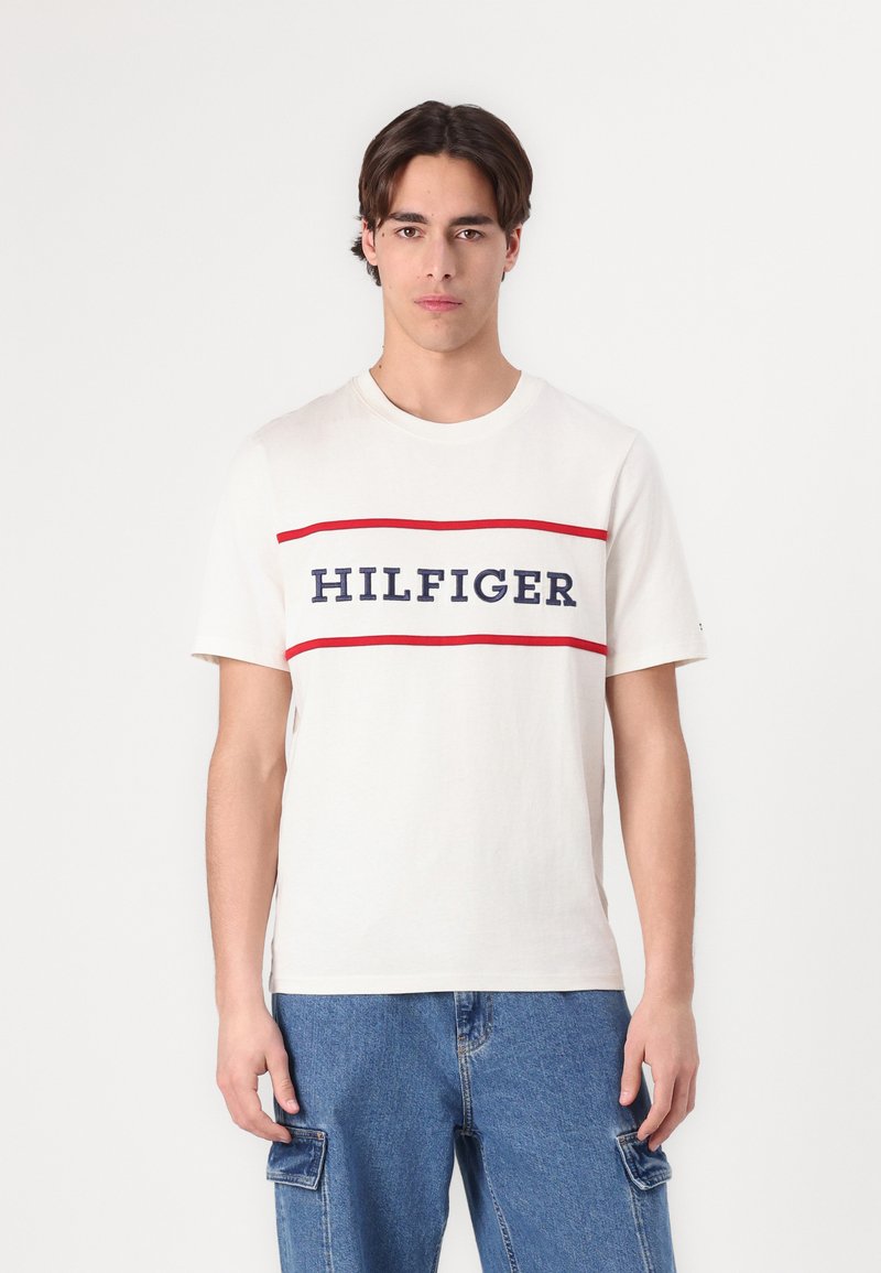 White cotton t-shirt with round neck, featuring a central navy and red 'HILFIGER' logo across the chest, worn with blue cargo shorts.