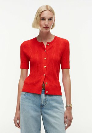 Femme aux cheveux blonds courts portant un cardigan rouge à manches courtes boutonné et un jean bleu clair, debout devant un fond blanc.