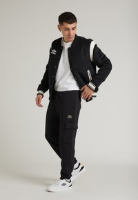 Giubbotto varsity nero con dettagli bianchi, abbinato a pantaloni cargo scuri con tasche, e sneakers bianche con dettagli neri e gialli.