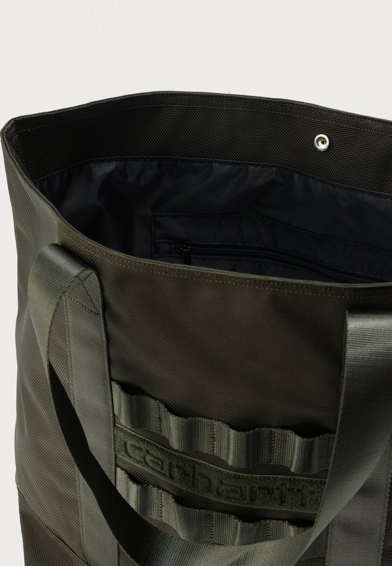 Dark green Carhartt tote bag with wide handles, external webbing detail, and open top showing black interior with zippered pocket.