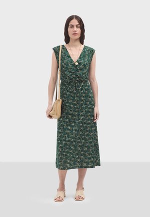 Drape jurk in groene stof met een geometrisch patroon, diepe V-halslijn, cinched taille en kapmouwen, gecombineerd met beige accessoires.