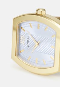 Relógio em tom de ouro com caixa retangular, mostrador texturizado azul claro, marcadores de horas dourados e bracelete texturizado. Apresenta o nome da marca "Guess" às 12 horas.