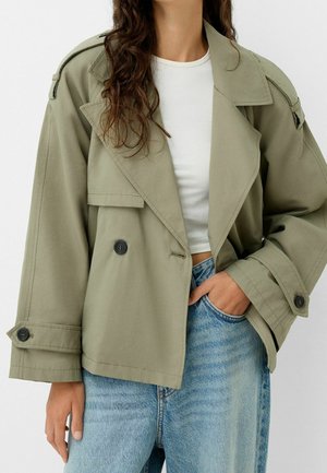 Donna che indossa una giacca doppiopetto verde oliva oversize, un top bianco corto e jeans a vita alta azzurri con una mano in tasca.