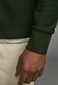 Grünes gestricktes Langarmshirt aus strukturiertem Stoff. Manschette hochgerollt, über einer hellgrauen Schicht getragen. Hand ruht auf einem hellen Hintergrund.