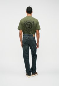 Homem em T-shirt gráfica verde-oliva, jeans azul-escuro e ténis castanhos, de pé voltado para longe contra um fundo branco liso.