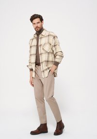 Beżowa koszula w kratę z brązowymi akcentami, w połączeniu z brązowym golfem, beżowymi spodniami i ciemnobrązowymi butami zapinanymi na sznurowadła.