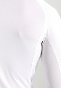 Balts sporta krekls ar teksturētu, elpojošu režģa dizainu. Iespējamās cieši pieguļošas piedurknes un smalks savilkuma details plecu šuvē.