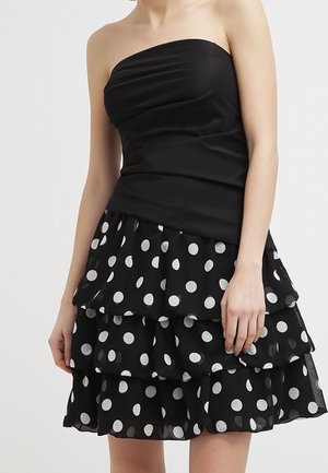 Schwarzes trägerloses Kleid mit einem mehrlagigen Rock mit weißen Punkten, getragen von einer Person mit entspannten Armen seitlich am Körper.