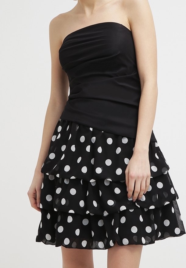 Robe noire sans bretelles avec une jupe superposée ornée de pois blancs, portée par une personne les bras détendus le long du corps.