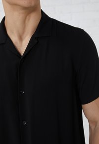 Man wearing a black short-sleeve button-up shirt with a spread collar, cropped from the neck to mid-torso.