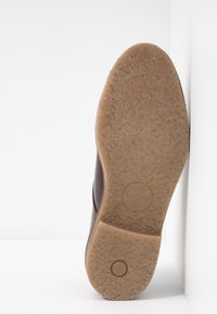 Braune Lederschuhe mit einer strukturierten Gummisohle, die ein rundes Logo und einen glatten, leicht abgerundeten Absatz aufweisen.