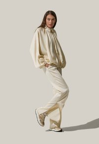 Blusa oversize color crema con fronte plissettato, abbinata a pantaloni su misura coordinati e sneakers beige con un design riflettente.