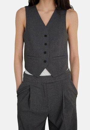 Femme portant un gilet boutonné gris foncé et un pantalon plissé taille haute assorti, les mains dans les poches.