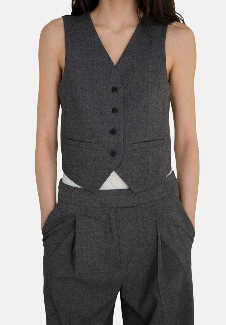 Femme portant un gilet boutonné gris foncé et un pantalon plissé taille haute assorti, les mains dans les poches.