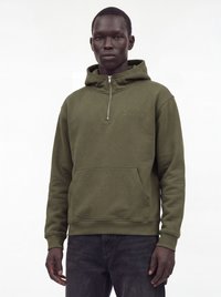 Homme portant un sweat à capuche vert olive à demi-fermeture éclair avec poche avant et pantalon sombre, debout devant un fond blanc uni.