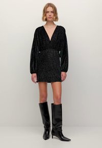 Czarna błyszcząca mini sukienka z głębokim dekoltem w serek i długimi rękawami, taliowana, w połączeniu z czarnymi kozakami na obcasie za kolano. Gładka faktura.