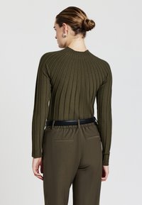 Maglione a coste verde oliva con collo alto, abbinato a pantaloni a vita alta coordinati e una cintura nera, che mostrano una silhouette aderente.