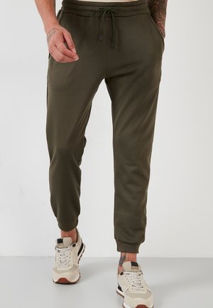 Mann trägt olivgrüne Jogginghosen mit Kordelzug an der Taille und weiße Turnschuhe, steht vor einem schlichten hellen Hintergrund.