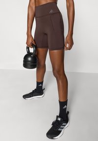 Καφέ σορτς ψηλόμεσα, ελαστικό ύφασμα, με μικρή τσέπη. Μαύρα αθλητικά παπούτσια με λευκές λεπτομέρειες και λογότυπο, συνδυασμένα με μαύρες κάλτσες.