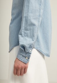Lyseblå denimskjorte med buet kant, knapmanchetter og subtile sydetaljer. Materialet synes blødt med en let struktureret overflade.