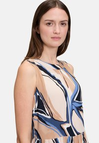 Ärmelloses Kleid aus fließendem Stoff, mit einem beige Grundton und abstrakten blauen und schwarzen Mustern, sowie einem runden Ausschnitt mit einer Schlüsselloch-Detail.