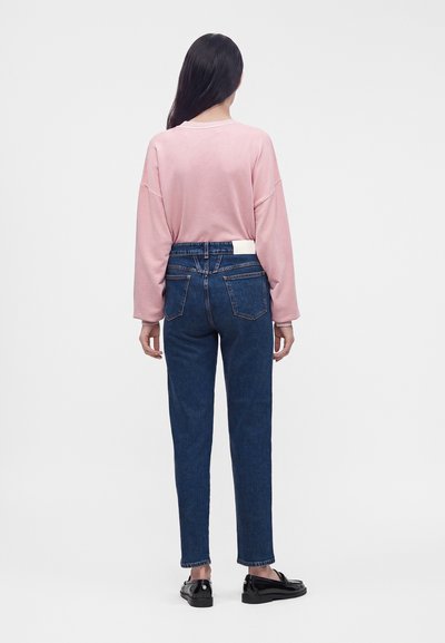 Femme aux longs cheveux foncés, vue de dos, portant une chemise rose à manches longues, un jean bleu et des mocassins noirs sur un fond uni.
