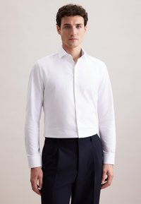 Man in een aansluitend wit overhemd met knopen en een zwarte broek, staande tegen een effen lichte achtergrond, recht van voren kijkend.