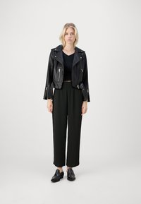 Svart lær cropped bikerjakke med glidelåser, kombinert med svarte vide bukser og svarte sko. En ensfarget bakgrunn forbedrer synligheten.