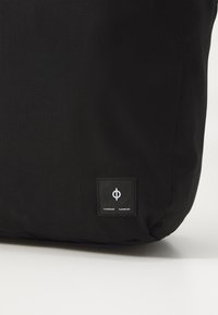 Svart veske laget av teksturert materiale, med et kvadratisk merke som har et hvitt emblem og merkenavn. Designet er enkelt og moderne.